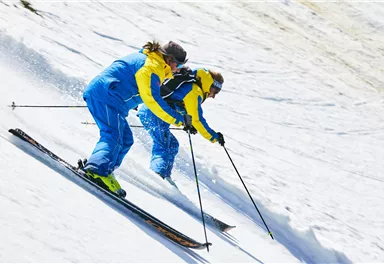 Two female skiers in blue and yellow functional clothing are skiing down a snow-covered slope together. The clear sky and fresh layer of snow create a sporty atmosphere.