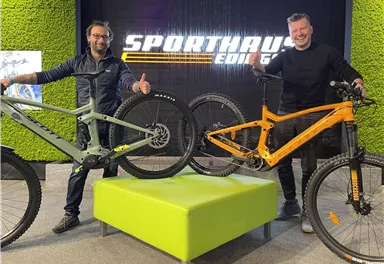 Zwei Männer stehen neben modernen Mountainbikes in einem Geschäft. Der Hintergrund zeigt den Namen „Sporthaus“ und eine grüne Wand.
