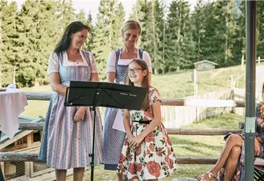Drei Frauen in traditioneller Tracht stehen auf einer Terrasse. Ein Mädchen lächelt und hält ein Papier für eine Aufführung.