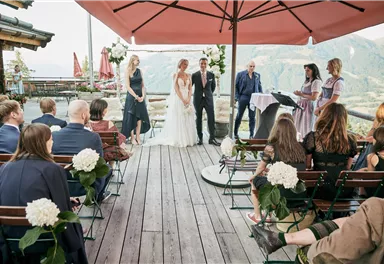 Eine romantische Hochzeit im Freien mit Blick auf die Berge. Braut und Bräutigam stehen im Mittelpunkt, während die Gäste auf Bänken sitzen.