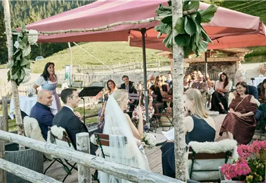 Eine romantische Hochzeitszeremonie im Freien unter einem Pavillon. Umgeben von Gästen, die gebannt der Braut und dem Bräutigam lauschen.