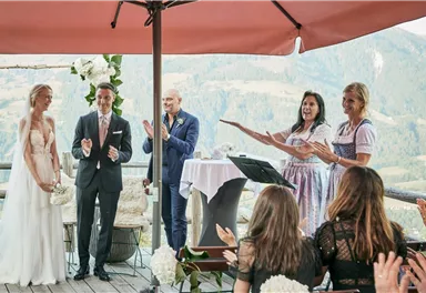 Eine Hochzeit im Freien mit magischer Berglandschaft im Hintergrund. Das Brautpaar steht vor Gästen und einer Zeremonie, umgeben von Blumen und festlicher Stimmung.