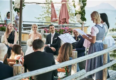 Eine wunderschöne Hochzeit im Freien mit Bergen im Hintergrund. Die Braut und der Bräutigam sitzen umgeben von Gästen, während das Zeremonienmeister vorliest.