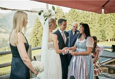 Eine Hochzeit im Freien mit einer fröhlichen Atmosphäre. Das Paar tauscht Ringe, umgeben von Familie und Freunden in einer malerischen Berglandschaft.