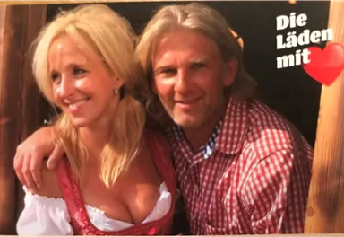 Ein lächelndes Paar steht zusammen vor einem Holzgebäude. Die Frau trägt ein traditionelles bayerisches Dirndl und der Mann ein kariertes Hemd.
