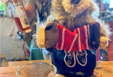 Eine lustige Dekofigur eines Murmeltieres mit traditioneller Kleidung steht auf einem Tisch. Im Hintergrund hält eine Person eine Flasche in der Hand und bereitet sich auf trinken vor.