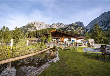 Eine malerische alpenländische Hütte mit einem schönen Garten und Blick auf beeindruckende Berge. Die Umgebung ist grün und ein ruhiger Bach fließt vorbei.