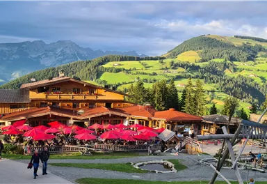 Ein gemütliches Restaurant mit rotem Sonnenschutz in einer malerischen Berglandschaft. Die Umgebung ist grün und hügelig, umgeben von Bergen und einem klaren Himmel.