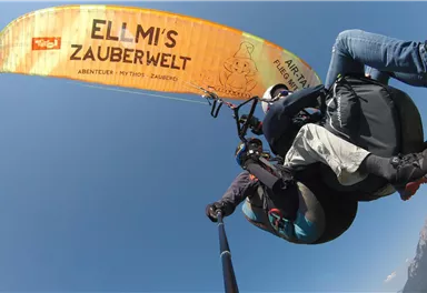 Ein Paragliding-Flug über eine malerische Landschaft mit blauer Himmel. Der Gleitschirm trägt den Schriftzug "ELLMI'S ZAUBERWELT".
