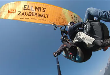 Ein Paragliding-Flug über eine malerische Landschaft mit blauer Himmel. Der Gleitschirm trägt den Schriftzug "ELLMI'S ZAUBERWELT".