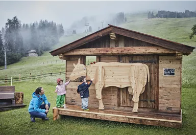 Ein Holzhaus mit einer Holzfigur einer Kuh steht auf einer Wiese. Zwei Kinder und eine Frau stehen davor und erkunden die Figur.