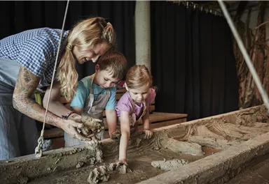 Eine Frau zeigt zwei Kinder, wie man mit Lehm arbeitet. Sie formen gemeinsam etwas in einer Werkstatt.