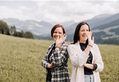 Two women are standing in a meadow, smiling and waving at the camera. In the background, green hills and mountains are visible.