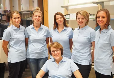 A group of six people in light blue shirts stands together in a room. One person is sitting in the foreground and smiling kindly.