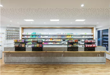 Ein modern gestalteter Apothekeninnerer mit Regalen voller Medikamente. Der Empfangstresen aus Beton steht im Vordergrund und bietet eine freundliche Atmosphäre.