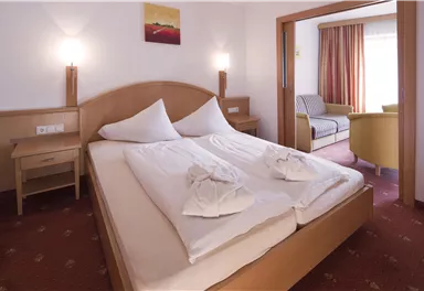 A cozy bedroom with a large double bed and bright bedding. In the background, there is a comfortable seating area and plenty of daylight.
