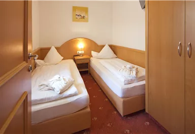 A simple room with two single beds, warm wooden furniture, and a wardrobe. A small picture hangs on the wall, and there is cozy lighting.