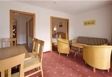 A cozy living room with a dining table and several chairs. The room is bright and modernly furnished, with a sofa and an adjacent bedroom.