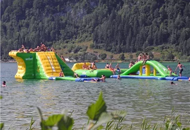 Ein großes, grünes Wasser-Spielgerät steht im See, umgeben von Bäumen. Viele Menschen haben Spaß beim Springen und Rutschen.