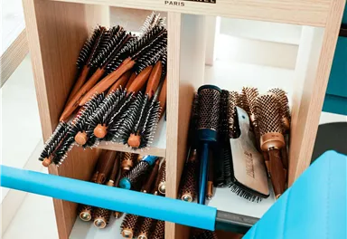 Ein Holzregal mit verschiedenen Haartools und -bürsten. Oben liegt ein schwarzes Gürtelset zur Aufbewahrung von Werkzeugen.