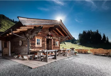 Eine rustikale Holzhütte in einer malerischen Berglandschaft. Der Himmel ist klar und die Sonne scheint, während die Terrasse einladend aussieht.