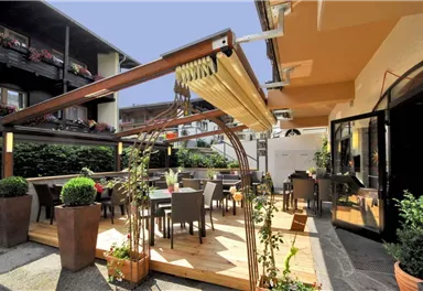 Ein gemütlicher Außenbereich eines Restaurants mit Holzterrasse und Tischen. Umgeben von Pflanzen und einem klaren Himmel.