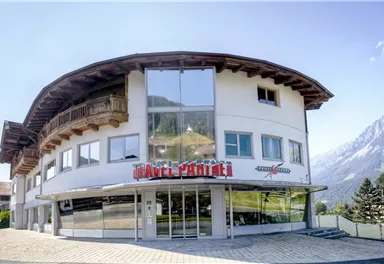 Ein modernes Gebäude mit großen Fenstern und einem geschwungenen Dach. Im Hintergrund sind Berge und ein klarer Himmel zu sehen.