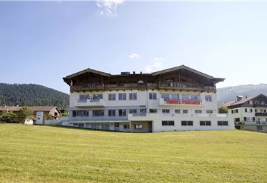 Ein modernes Hotelgebäude mit einem traditionellen Dach in einer grünen Wiesenlandschaft. Im Hintergrund sind sanfte Hügel und ein blauer Himmel zu sehen.