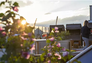 A beautiful rooftop terrace with blooming plants in the foreground and a stunning sunset in the background. A person stands nearby and enjoys the view.