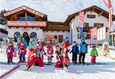 A ski school with a colorful building in the background. In front of the building, ski instructors and children in ski clothing are standing on snow.