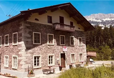 A charming stone building with a spacious balcony. In the background, mountains and a blue sky can be seen.