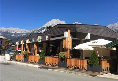A cozy restaurant with a wooden terrace and sun umbrellas. Majestic mountains and a blue sky can be seen in the background.