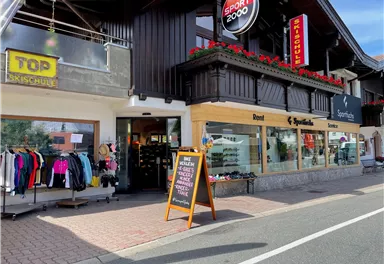 A sports store with a large selection of sports clothing and equipment. The facade is decorated with flowers, and there is a sign indicating special offers.