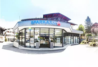 Eine moderne Bankfiliale mit großen Fenstern und einem einladenden Eingangsbereich. Sie befindet sich in einer ruhigen Umgebung mit Bäumen und Gebäuden im Hintergrund.