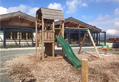 Ein Spielplatz mit einem Holz-Spielturm, einer grünen Rutsche und Schaukeln. Im Hintergrund sind moderne Gebäude mit großen Fenstern zu sehen.