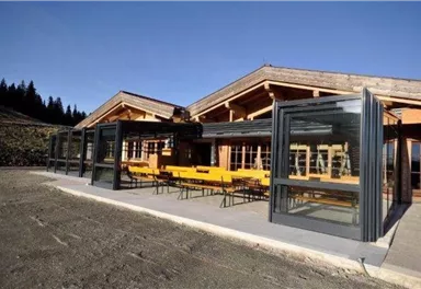 Ein modernes Gebäude mit großen Fenstern und einer Terrasse. Auf der Terrasse stehen gelbe Tische und Bänke.