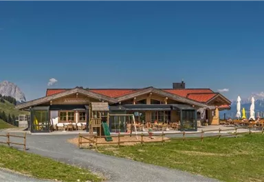 Ein gemütliches Bergrestaurant mit einer großen Terrasse. Im Hintergrund sind die Alpen und ein klarer blauer Himmel zu sehen.