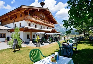 A charming wooden building with an inviting outdoor seating area.
Tables and chairs are on the lawn, surrounded by mountains and a blue sky.