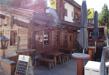 A rustic cabin with wooden cladding and an outdoor area. There are tables, umbrellas, and a sign that reads "Top Bar."