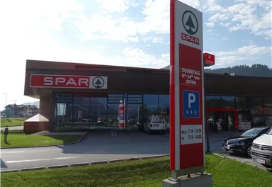 A SPAR supermarket with a large parking lot and clear blue sky. The opening hours are indicated on the sign.