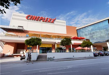 A modern cinema with the inscription "Cineplexx" on the facade. In front of the building, people and plants can be seen.