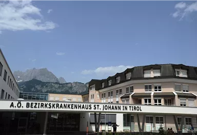 Ein modernes Krankenhausgebäude mit dem Namen "A.O. Bezirkskrankenhaus St. Johann in Tirol". Im Hintergrund sind beeindruckende Berge zu sehen.