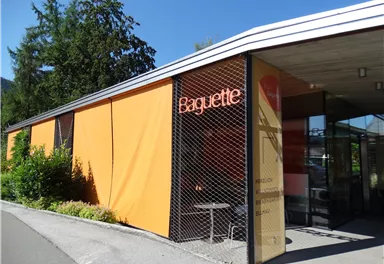 A modern building with a striking orange facade and the inscription "Baguette". Surrounded by green plants and under a clear blue sky.