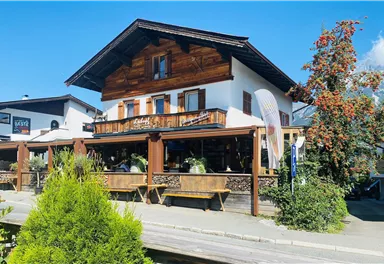 A charming wooden house with an inviting terrace. Surrounded by plants and a blue sky.