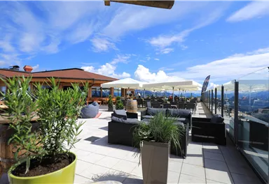 A bright rooftop terrace with comfortable seating and umbrellas. In the background, a blue sky with a few clouds is visible.