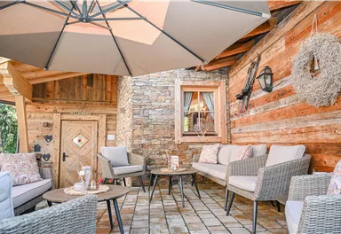 A cozy seating area with comfortable furniture under a large sunshade. The wall is made of beautiful wood and stone, creating an inviting atmosphere.