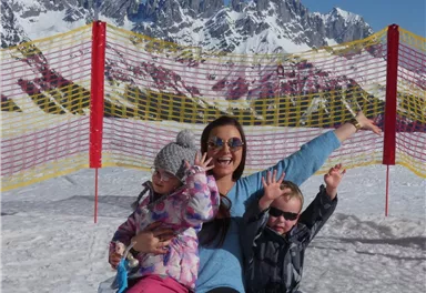 A cheerful woman is sitting in the snow with two children and posing. Snow-covered mountains can be seen in the background.