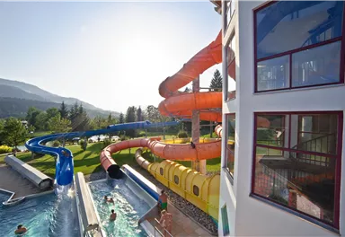 Ein Wasserpark mit mehreren Rutschen und einem Schwimmbecken. Die Sonne scheint, und die Umgebung ist grün und einladend.