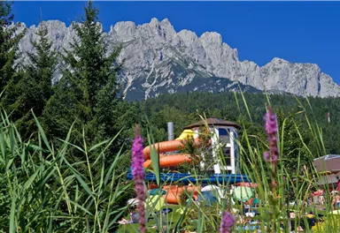 Ein Wasserpark mit bunten Rutschen und grünen Wiesen. Im Hintergrund sind majestätische Berge und ein klarer blauer Himmel zu sehen.