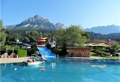 Ein schöner Pool mit einer Wasserrutsche und Blick auf die Berge. Umgeben von grünen Bäumen und sonnigem Wetter.